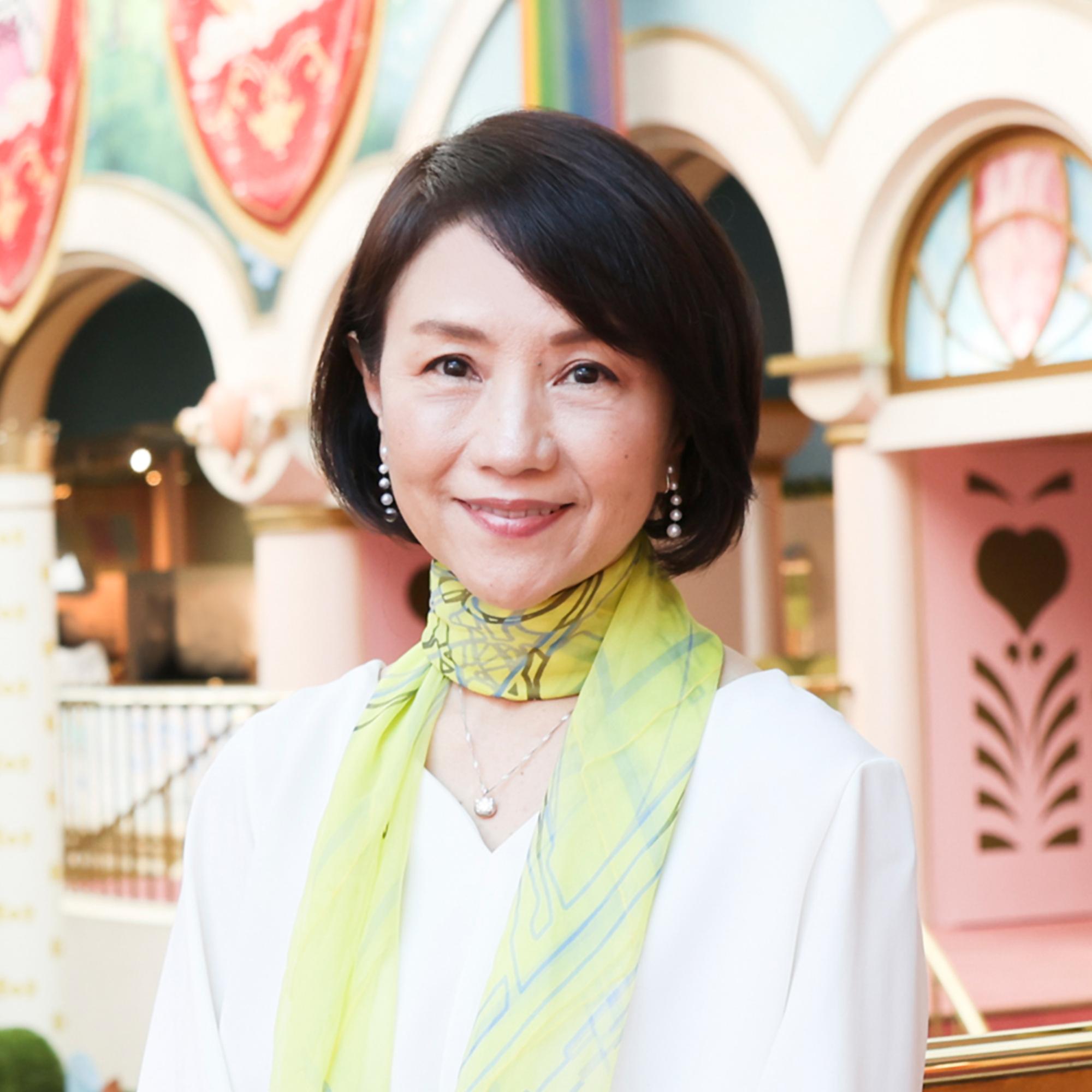 白いトップスと黄緑色のスカーフを身に纏い、穏やかな微笑みを浮かべて正面を見つめる小巻亜矢さんの写真