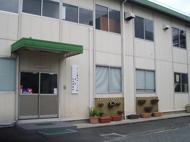 薄いベージュ色の外壁に緑色の庇が玄関に設置された2階建ての建物で、入り口横には「エース構内サービス株式会社 本社 九州事業所」と書かれた白い看板が掲げられ、窓の下にはプランターや木製のラティスが飾られている写真
