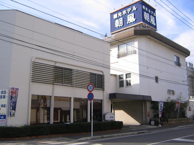 屋上に「観光ホテル 朝風」と大きく書かれた青い看板を掲げた白い外壁の三階建ての建物が、道路沿いに建っており、歩道側には低い生垣や標識、宴会の案内などののぼり旗が並んでいる建物外観の写真