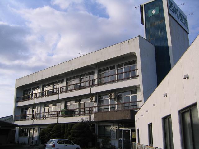屋上付近の青い塔屋部分には緑色の十字マークと「西村医院」という大きな看板が掲げられており、白い外壁の3階建ての西村医院の外観写真