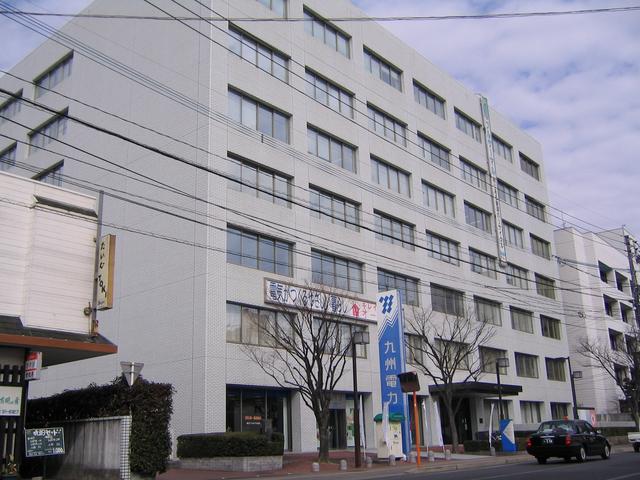 薄いグレーのタイルが整然と並ぶ多層階の大規模なオフィスビルで、歩道沿いには「九州電力」と書かれた青い縦型の看板が立つ九州電力株式会社 佐賀支社の外観写真