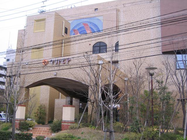 ベージュや薄茶色のタイルを基調とした大規模な建物で、大きなアーチ型の車寄せや上部に描かれた円形の青いアートパネルが目を引き、建物の前面には多くの木々が植えられ「マリトピア」というオレンジ色のロゴが掲げられている写真