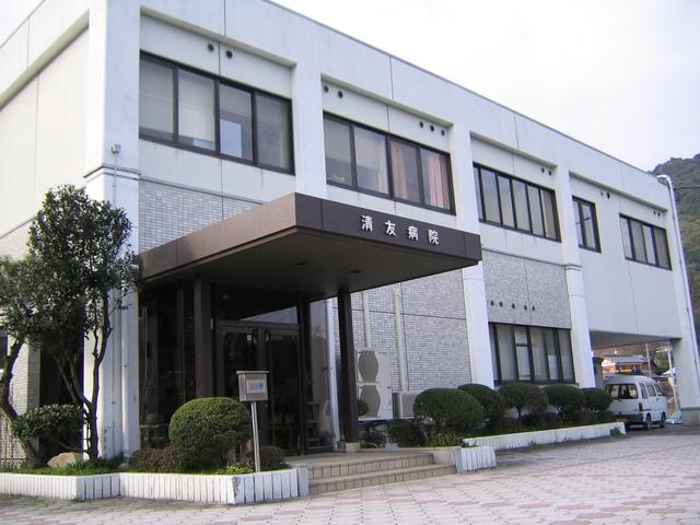 建物前には丸く整えられた植栽が並び、白を基調とした外壁に整然と窓が並ぶ2階建ての建物で、正面入り口の茶色い大きなひさし部分に白文字で「清友病院」と記された、清友病院の外観写真