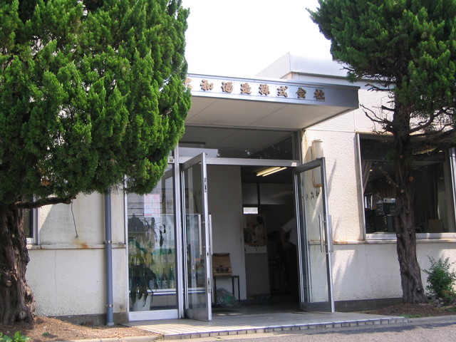銀色の庇の正面に「大和酒造株式会社」と黒い文字で社名が記されており、大きく開かれたガラスの二枚扉の向こうに店内の様子が見え、左右を立派な常緑樹の庭木に囲まれた事務所エントランスの写真