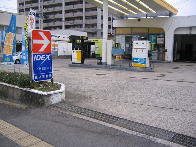 道路沿いに設置された「IDEX 新出光グループ 理研石油株式会社」と書かれた青と赤の看板の奥に、給油機や洗車設備のあるガソリンスタンドの写真
