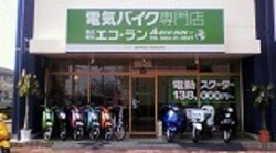 「電気バイク専門店 株式会社エコ・ラン」という緑色の大きな看板が掲げられ、店頭には色鮮やかな電動スクーターが並び、ガラス窓には「電動スクーター 138,000円〜」という案内が貼られた店舗の外観を捉えた写真