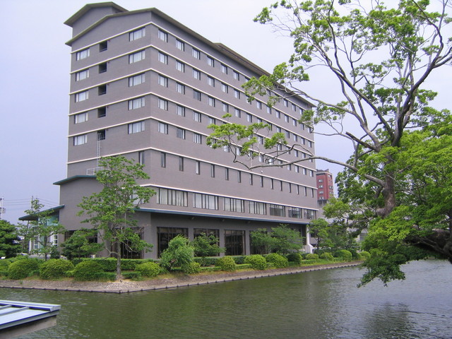 静かな水面に面して建つグレーの外壁が落ち着いた印象の多階建てホテルで、各階に並ぶ窓のラインが美しく、建物の周囲には手入れされた低い生け垣や木々が配され、水辺の緑と建物が調和した景色が広がるホテルニューオータニ佐賀の全景写真