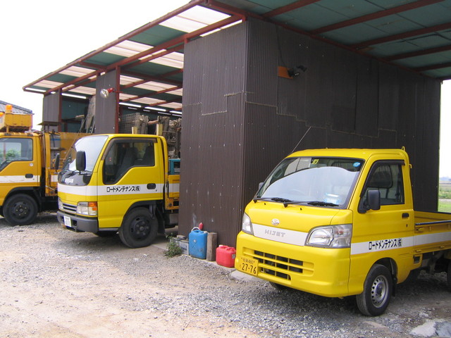 屋根付きの車庫に、側面に「ロードメンテナンス株式会社」と書かれた黄色いトラックと軽トラックが並んで停められている写真