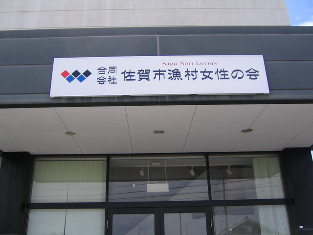 建物の入口上部に、青と赤の菱形ロゴとともに「合同会社 佐賀市漁村女性の会」と記された白地の看板が掲げられた外観写真