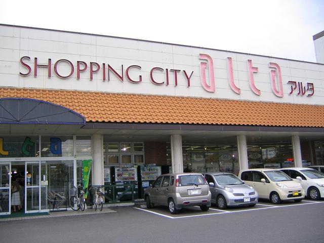 オレンジ色の瓦屋根が特徴的な白い外壁の建物に、赤く縁取られた「SHOPPING CITY alta アルタ」の大きな文字看板が掲げられ、店舗前には数台の乗用車が停車している、晴れた日のショッピングセンターの外観写真