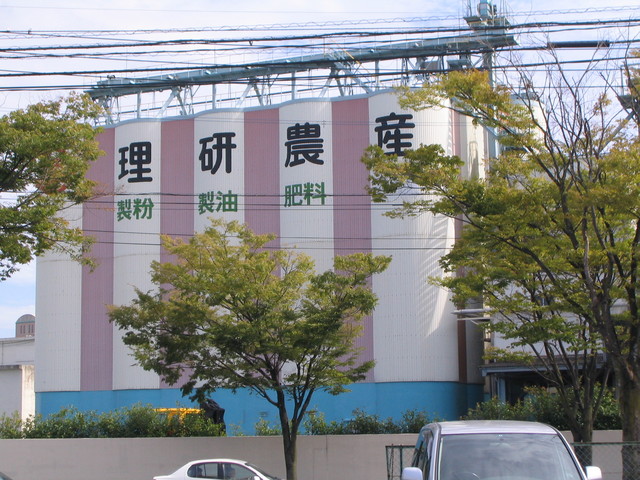 白とピンクの縦縞模様の巨大なサイロに「理研農産」と大きな黒文字、その下に緑色で「製粉・製油・肥料」と書かれた工場建屋が立ち並ぶ理研農産化工株式会社の写真