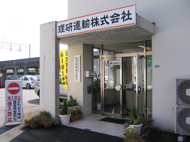 白い外壁の建物の入り口部分に、太い黒文字で「理研運輸株式会社」と大きく書かれた看板が掲げられてた理研運輸株式会社の外観写真