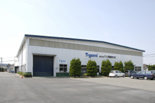 青空の下に佇む白い壁面の大きな工場建屋で、壁面には青色の「T-gami」という企業ロゴが掲げられ、手前の舗装されたスペースには複数の乗用車が駐車され、建物の前には等間隔に整えられた緑の植栽が並ぶ株式会社 戸上デンソーの外観写真