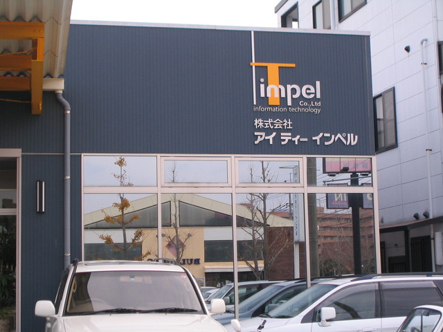 駐車場に車が数台停まっている奥に、濃紺の壁面にオレンジ色の大きなTと白文字のimpelが特徴的なロゴマークが掲げられ、その下に株式会社アイティーインペルという社名が白字で表記されている建物の外観写真