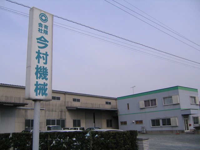 淡い紫色の空を背景に、左手前に大きな「有限会社 今村機械」と青い文字で書かれた縦長の白い看板が立ち、その奥にベージュ色の工場棟と、上部に緑色のラインが入った薄いグレーの2階建て事務所棟が並んでいる建物の外観写真