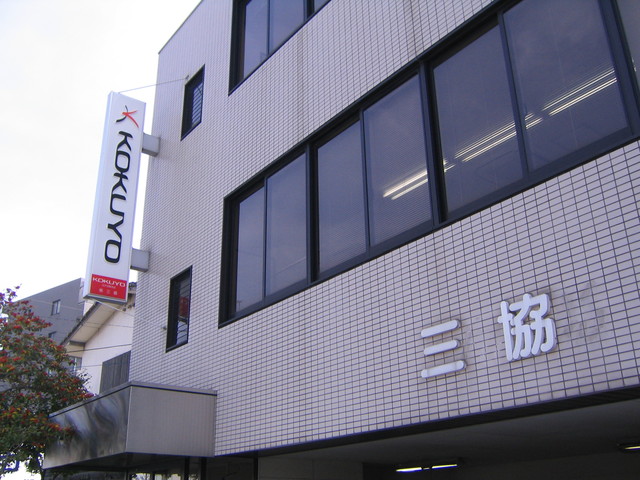 白いタイル貼りのビル壁面に「三協」という白い切り文字看板が設置され、その左側には「KOKUYO」のロゴが入った縦長のデザイン看板が突き出すように取り付けられている、オフィスビルの一部を斜め下から見上げた写真