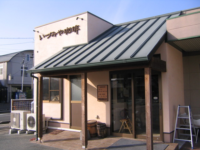 薄桃色の外壁に「いづみや珈琲」の店名が掲げられ、緑色の勾配屋根の庇が玄関ポーチを覆う落ち着いた佇まいの店舗外観と、入り口横に置かれた木樽や営業時間の案内板、店先に差し込む穏やかな日差しを捉えた写真