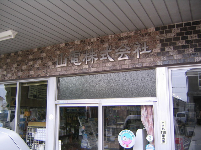 茶色や黒のモザイクタイルが装飾された外壁の入り口上部に、銀色の立体的な文字で「山電株式会社」と社名が掲げられており、ガラス扉越しに商品や棚が並ぶ店内の様子が見える山電株式会社の外観写真