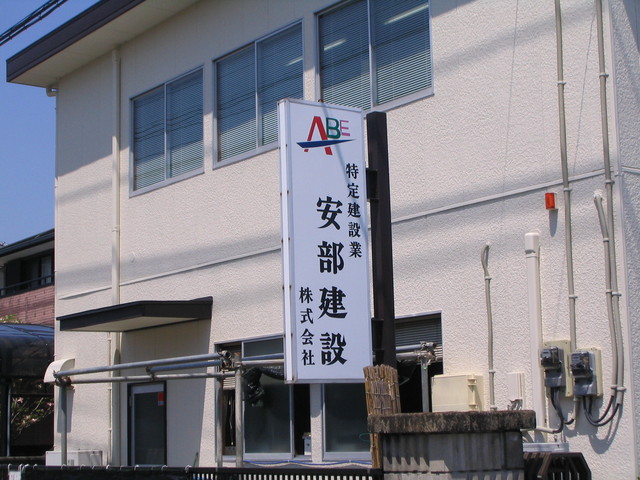 明るい日差しが注ぐ白い二階建ての建物の前に、赤と青の「ABE」のロゴマークと、黒い太文字で「特定建設業 安部建設 株式会社」と縦書きされた看板が設置されている建物外観の写真