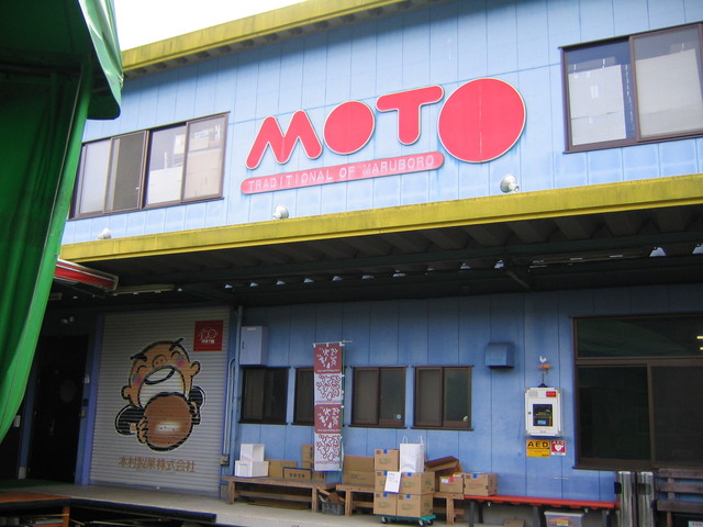 水色の外壁の建物に赤色で「MOTO」と大きく描かれた丸みのある看板が掲げられ、シャッターには丸い菓子を食べる子供のキャラクターと「本村製菓株式会社」の社名が記された、伝統的な丸ぼうろを製造する工場の写真