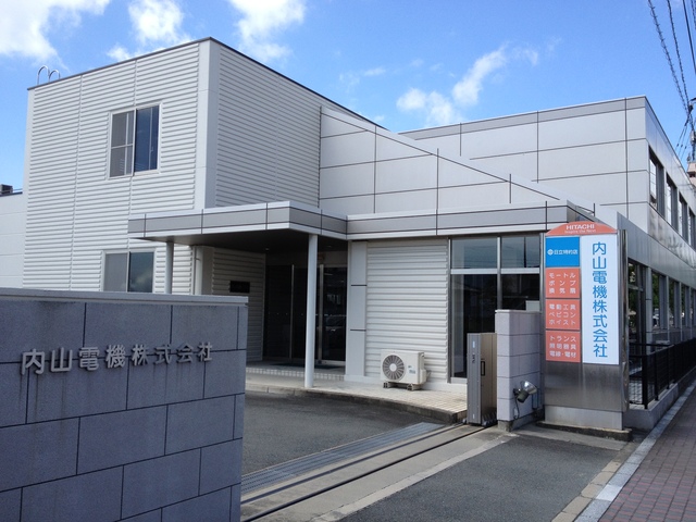 青空を背景に、白を基調としたスタイリッシュな外観の「内山電機株式会社」の社屋と、手前の門柱および右側の看板にそれぞれ刻印や掲示された社名、そして取り扱っている日立製品の品目が記された看板を捉えた写真