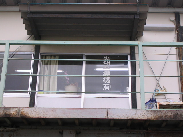 薄いベージュの外壁に小さな庇がついたプレハブ風の建物で、2階部分の窓ガラスには白い文字で「光武建機有限会社」と社名が記され、手前には淡い緑色の金属製手すりがあるベランダのようなスペースが写っている事務所の外観写真