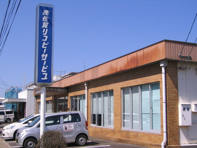 茶色のレンガ調の壁面が特徴的な平屋建ての事務所ビルの前に、「株式会社佐賀リコピーサービス」と白抜き文字で書かれた縦長の大きな青い看板が設置された佐賀リコピーサービスの外観写真