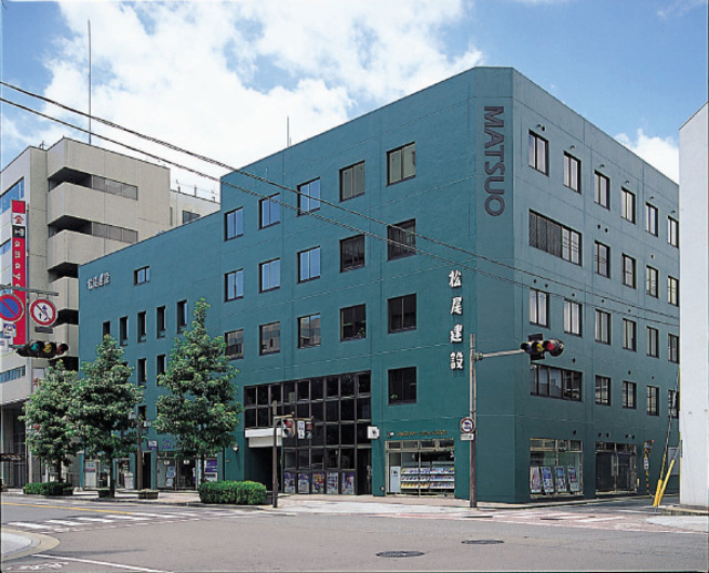 落ち着いた深緑色の外壁が特徴的な5階建てのビルで、壁面には「松尾建設」という社名と「MATSUO」の英字ロゴが大きく記され、1階のガラス張りの入り口付近には植栽が並ぶ、街角の交差点に面して建つ松尾建設ビルの外観写真