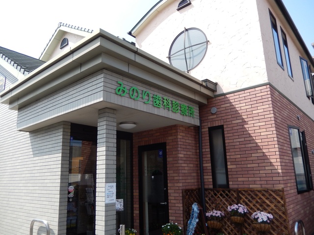 明るいベージュの塗り壁と赤茶色のレンガ風タイルが組み合わされた2階建ての建物の、みのり歯科診療所の外観写真