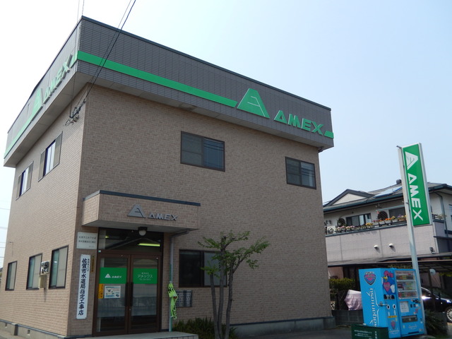 茶色のタイル貼りの二階建て社屋に、緑色の「AMEX」のロゴと社名が屋上付近のラインや玄関上、自立看板に統一して掲げられ、入り口横には「佐賀市水道局指定工事店」の看板が設置されている株式会社アメックスの外観写真