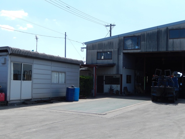 晴天の下、左側にプレハブ小屋、右側に開口部の大きなトタン壁の作業所が並び、その間の敷地に青いドラム缶や停車したトラックの後方が見える、静かな工場の敷地内を捉えた写真