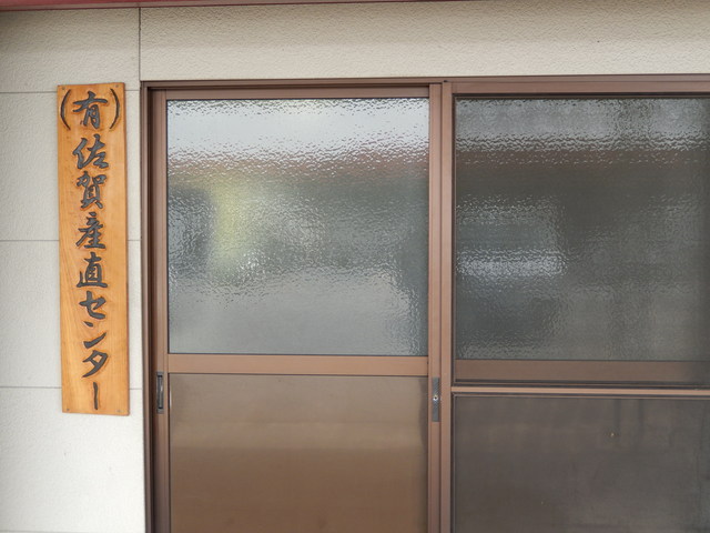 茶色のサッシの引き戸がある建物の入り口の左側に、「有限会社佐賀産直センター」と黒い文字で縦書きに彫られた木製の看板が掲げられた外観写真