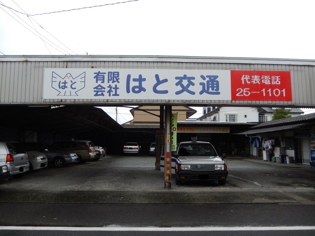 鳩のイラストロゴと共に「有限会社 はと交通」と大きく記された看板が掲げられ、平屋建ての有限会社はと交通の営業所の写真