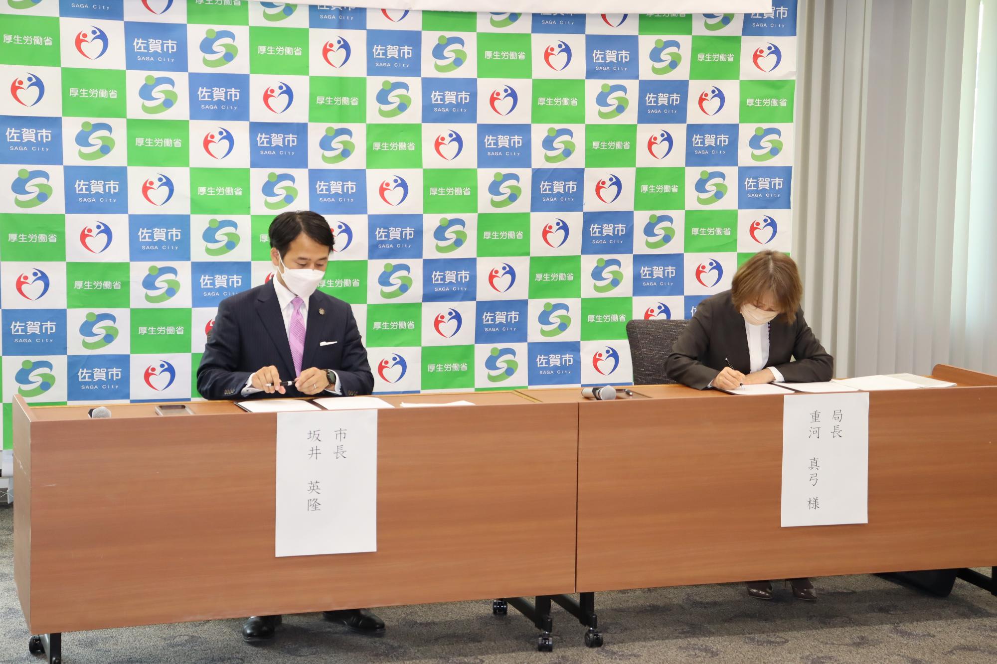 佐賀市と厚生労働省のロゴが並ぶバックパネルの前で、坂井英隆佐賀市長と重河真弓局長が並んで座り、木製の机に置かれた協定書等の書類にそれぞれ署名を行っている、厳粛な雰囲気の締結式の写真