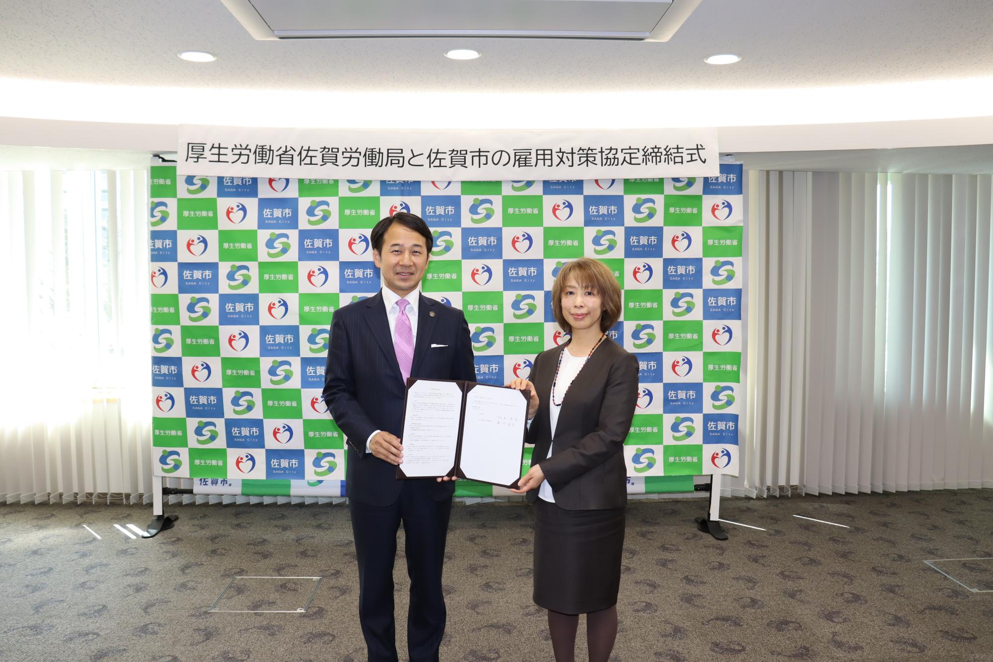 「厚生労働省佐賀労働局と佐賀市の雇用対策協定締結式」と書かれた横断幕が掲げられたバックパネルの前で、坂井英隆佐賀市長と重河真弓局長が、締結した協定書を広げて持ち、カメラに向かって並んで笑顔を見せている記念写真