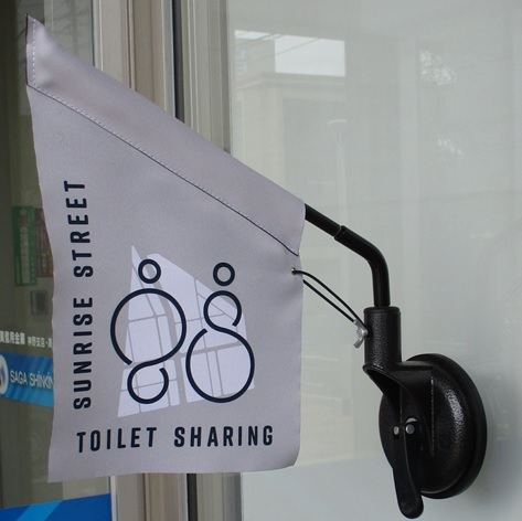 建物のガラス面に吸盤で固定されたグレーの小さな旗に、人の形を模したロゴマークと「SUNRISE STREET TOILET SHARING」という英字がプリントされ、街なかでのトイレ貸出協力中であることを示す写真