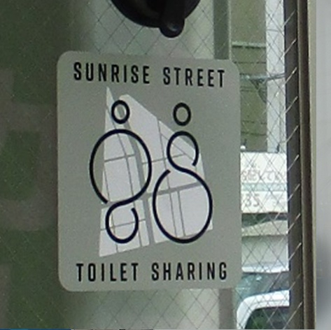 建物のガラス窓に貼られた四角いステッカーに、抽象的な二人の人物が並ぶロゴマークと「SUNRISE STREET TOILET SHARING」の英字が記され、街なかでトイレを共有する取り組みを示している写真