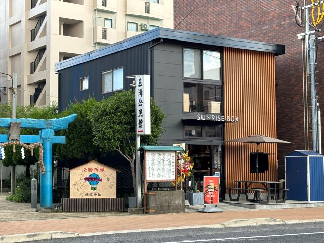 水色の鳥居や三溝公民館の看板の隣に立つ、JR佐賀駅近くのサンライズストリート沿いに位置する、黒い外壁と木目調のルーバーが特徴的な2階建ての建物「SUNRISE BOX」の建物外観写真
