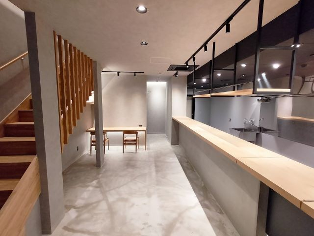 グレーを基調としたモダンな店内に、長い木製カウンターテーブルと奥の壁際に2脚の椅子が並ぶデスクスペース、左手には2階へ続く木目調の階段が設置された、シンプルで清潔感のある内装写真