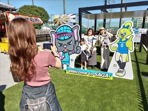 青空の下、人工芝が広がる屋外イベント会場で、佐賀のプロバスケットボールチームやバレーボールチームのキャラクターパネルを背にポーズをとる家族連れを、手前の女性がスマートフォンで撮影している微笑ましい風景の写真