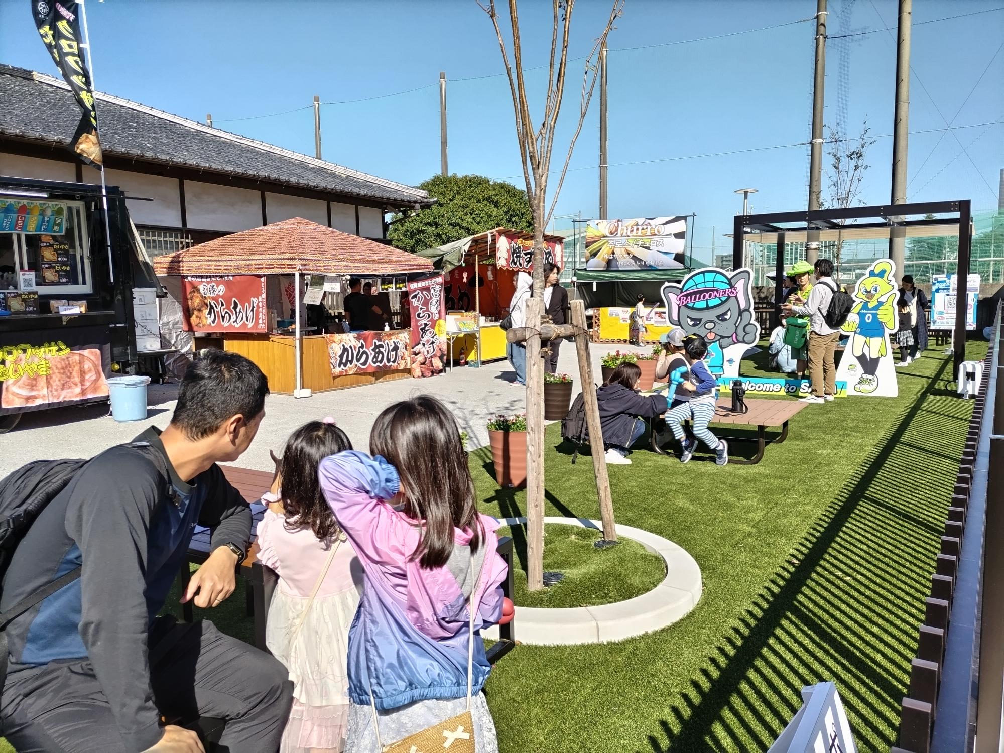 晴天の下、人工芝が敷かれた屋外広場で、唐揚げの屋台やキッチンカーが並び、マスコットキャラクターのパネルが設置された会場を、手前に座る親子連れが眺めている、活気あるイベント風景の写真