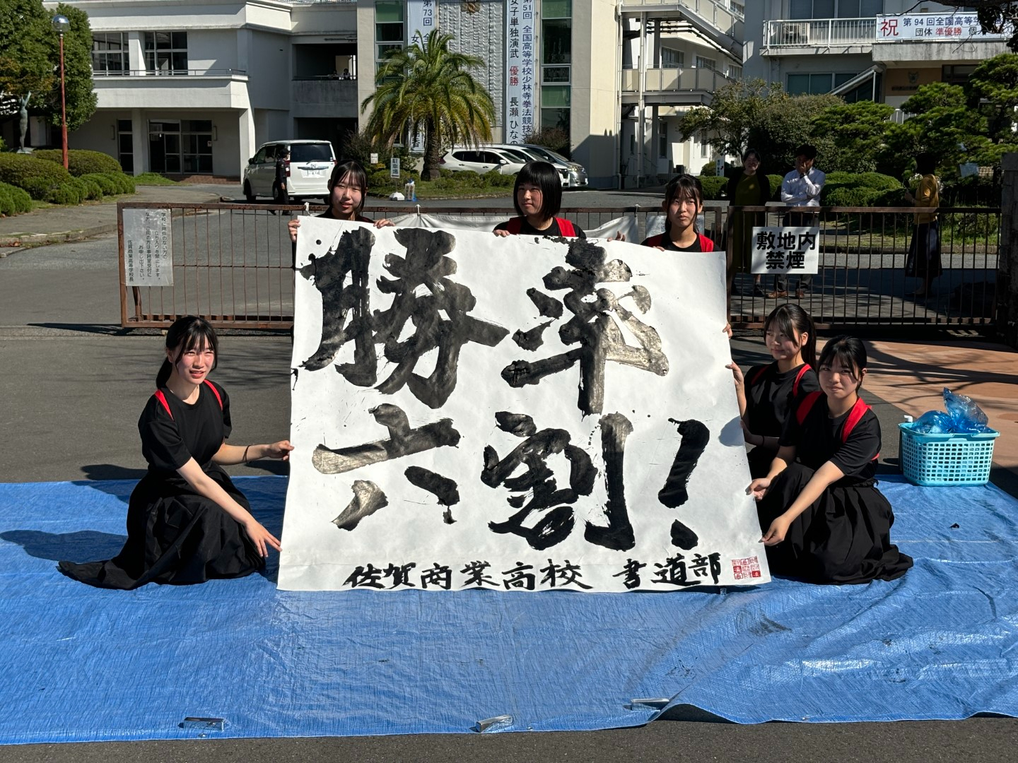 佐賀商業高校の校門前で、書道部の部員たちが「勝率六割！」と力強く書かれた大きな書を掲げ、青いシートの上に座って笑顔でポーズをとっている、晴天下の活気あふれる書道パフォーマンスを披露している様子の写真
