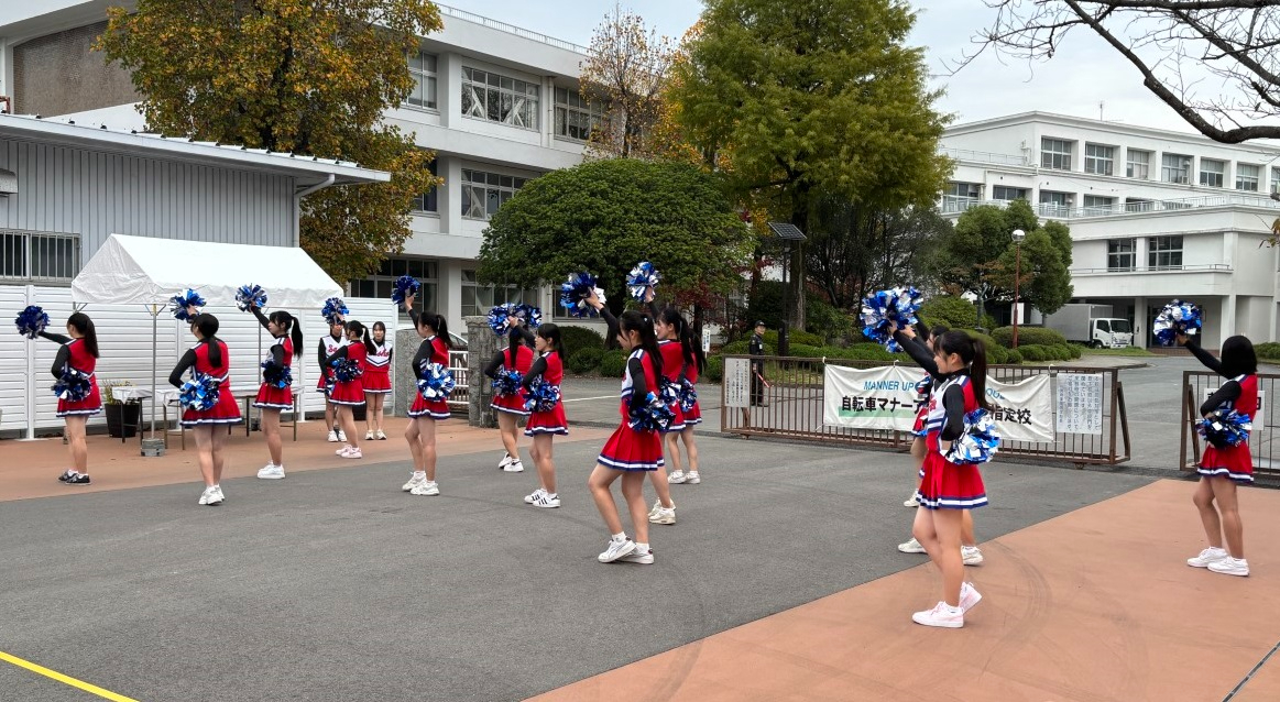 佐賀商業高校の校舎を背景にした屋外スペースで、赤と黒のユニフォームに身を包んだバトン部の生徒たちが、青と白のポンポンを掲げて華やかなパフォーマンスを披露している、活気に満ちたイベント風景の写真