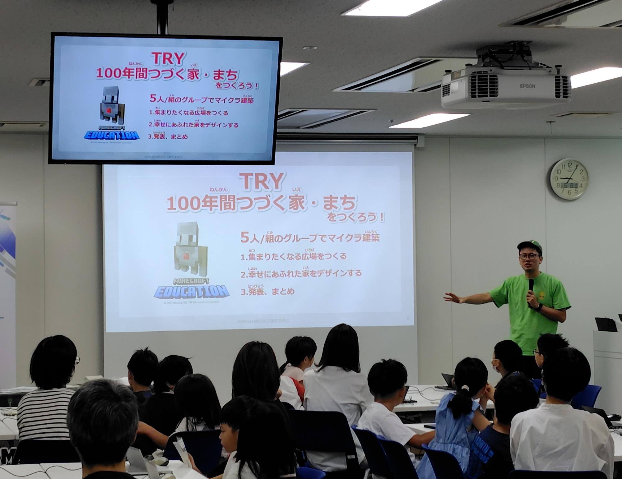明るい室内で「100年間つづく家・まちをつくろう！」というマインクラフトの教育プログラムのスライドを投影したスクリーンを前に、緑色のTシャツを着た男性が子供たちに身振り手振りを交えて説明をしている写真