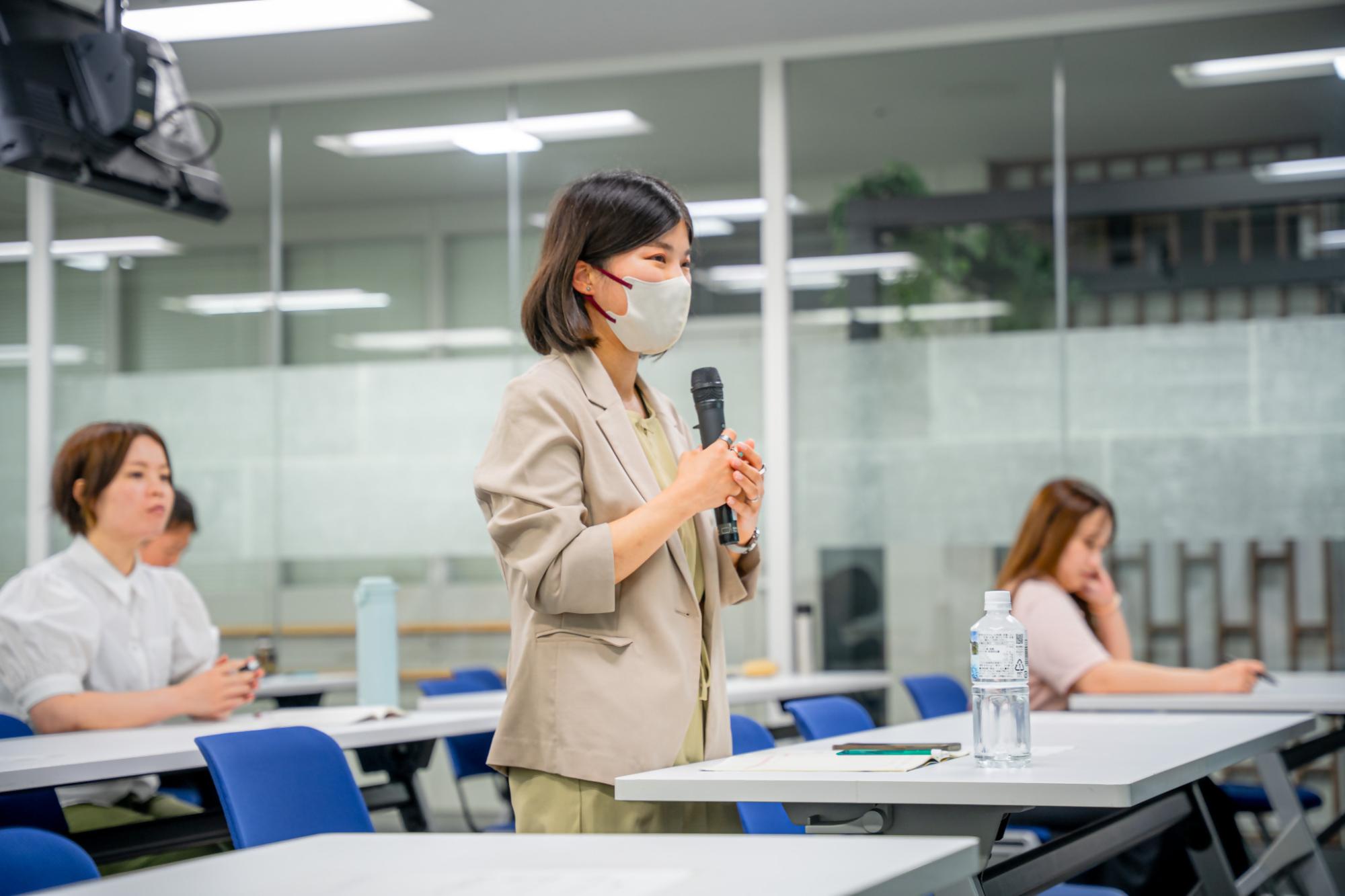研修会場で、女性がマイクを両手で持ち、他の受講生たちの前で立ち上がりながら質問や意見を述べている様子の写真