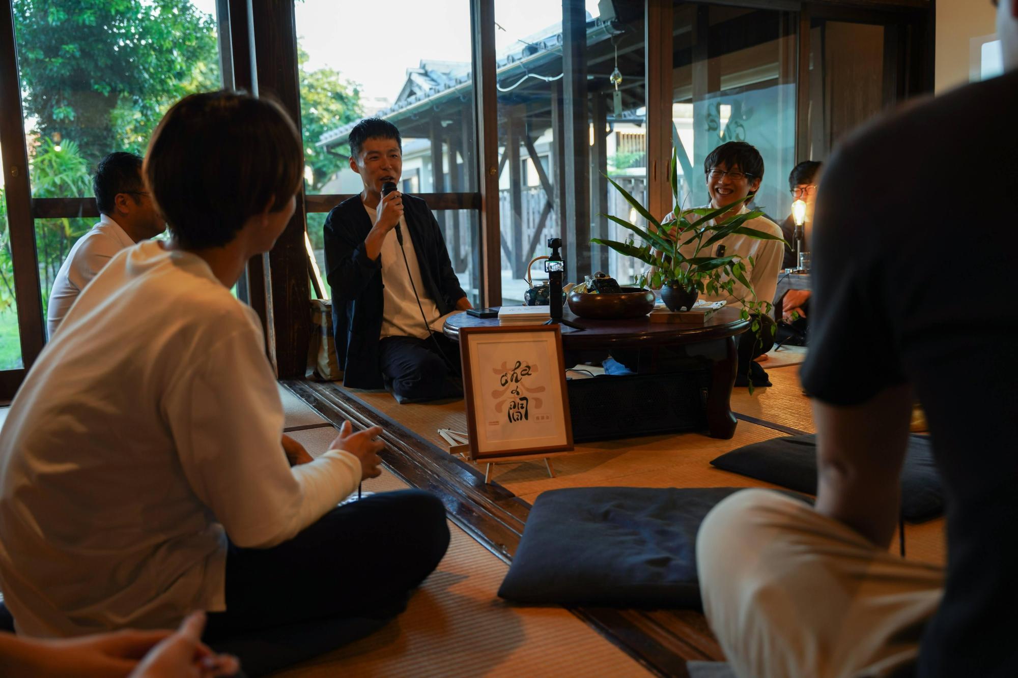 和風建築の畳の部屋で、中央の円卓を囲んで座る男性がマイクを手に笑顔で話し、それを見守る周囲の参加者とともに、室内から縁側越しに屋外の風景も見える落ち着いた和やかな座談会の様子を捉えた写真