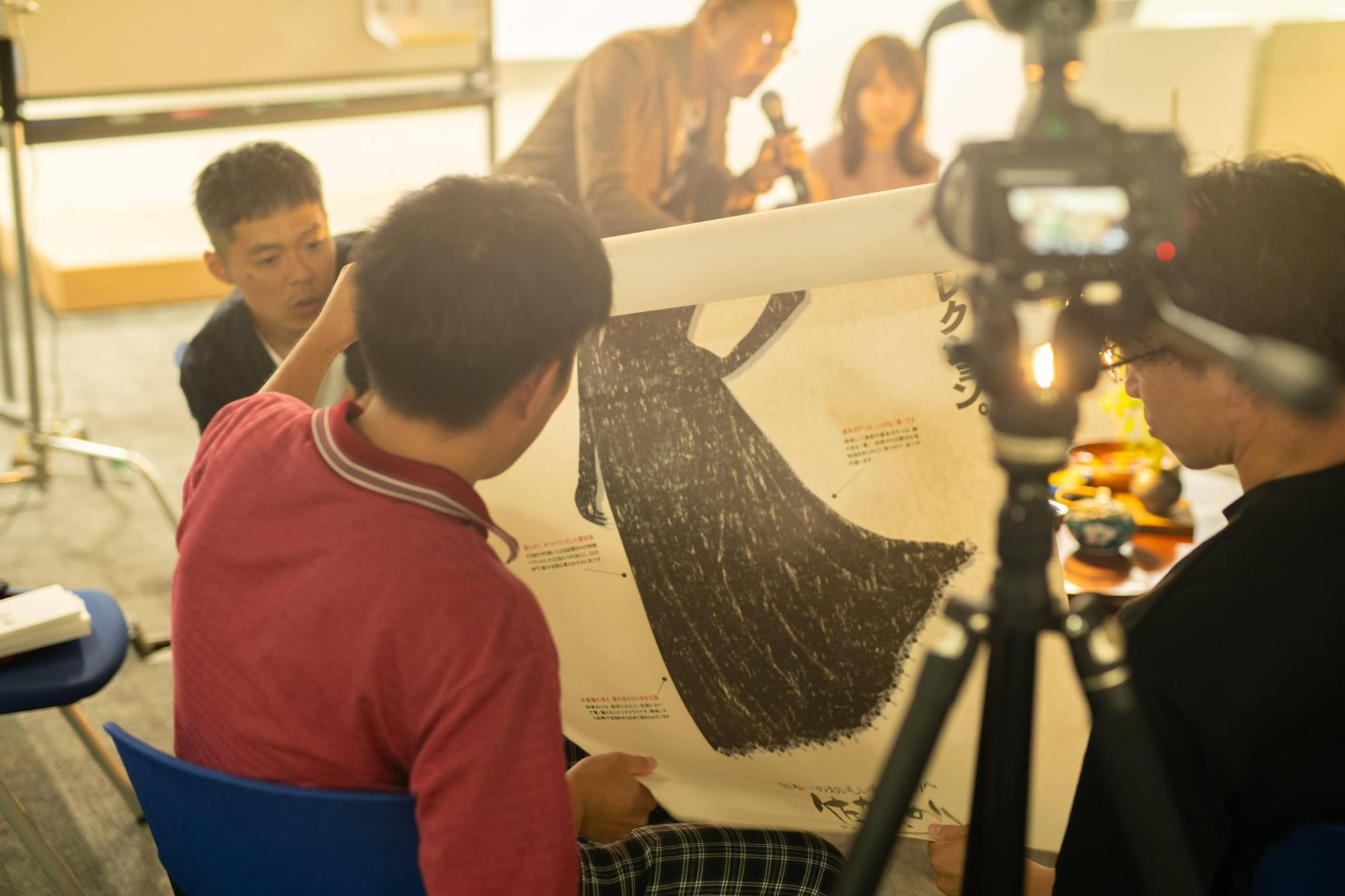 座談会会場にて、大きなイラストが描かれたポスター広げて真剣な表情で確認する参加者たちの姿を、手前の三脚に固定されたビデオカメラ越しに捉えることで、イベントの記録風景と現場の熱気を描写した写真