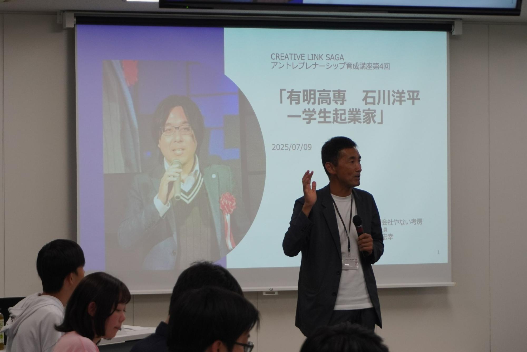 「アントレプレナーシップ育成講座第4回」のタイトルと石川洋平氏の顔写真が投影されたスクリーンの前で、マイクを握る講師が身振りを交えて解説を行い、熱心に耳を傾ける受講生たちの姿が手前に見える、講座の一場面を捉えた写真