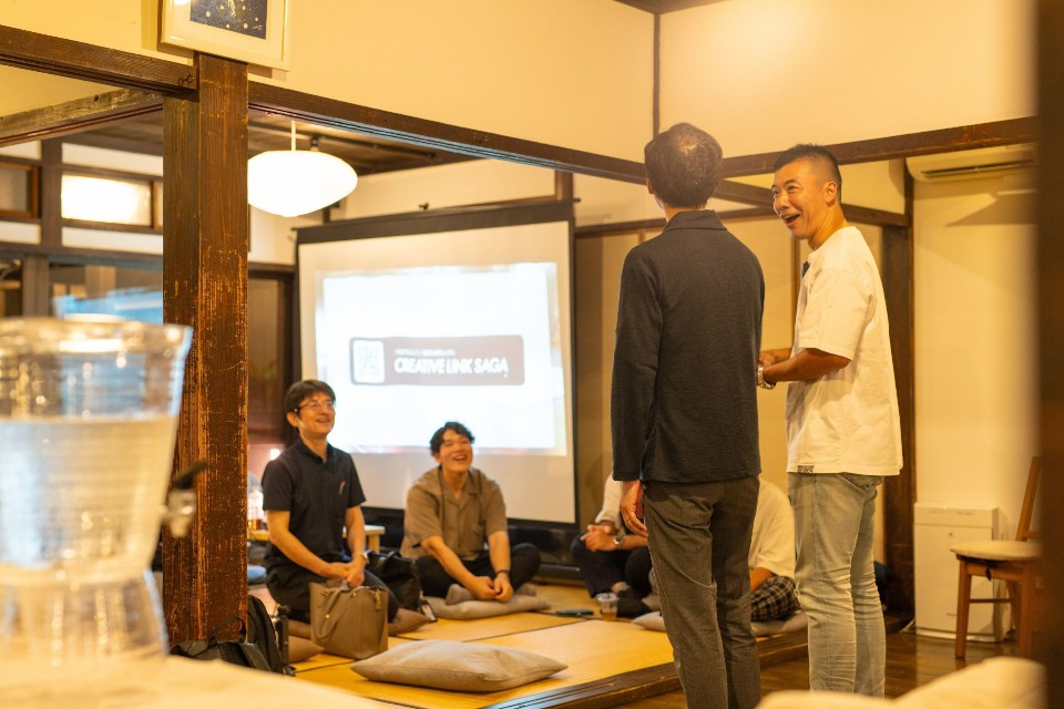 古民家風の広々とした和室で、前に立って話す2人の男性の漫才を見ている座布団に座りリラックスした様子で笑顔を浮かべる参加者たちを捉えた座談会の様子の写真