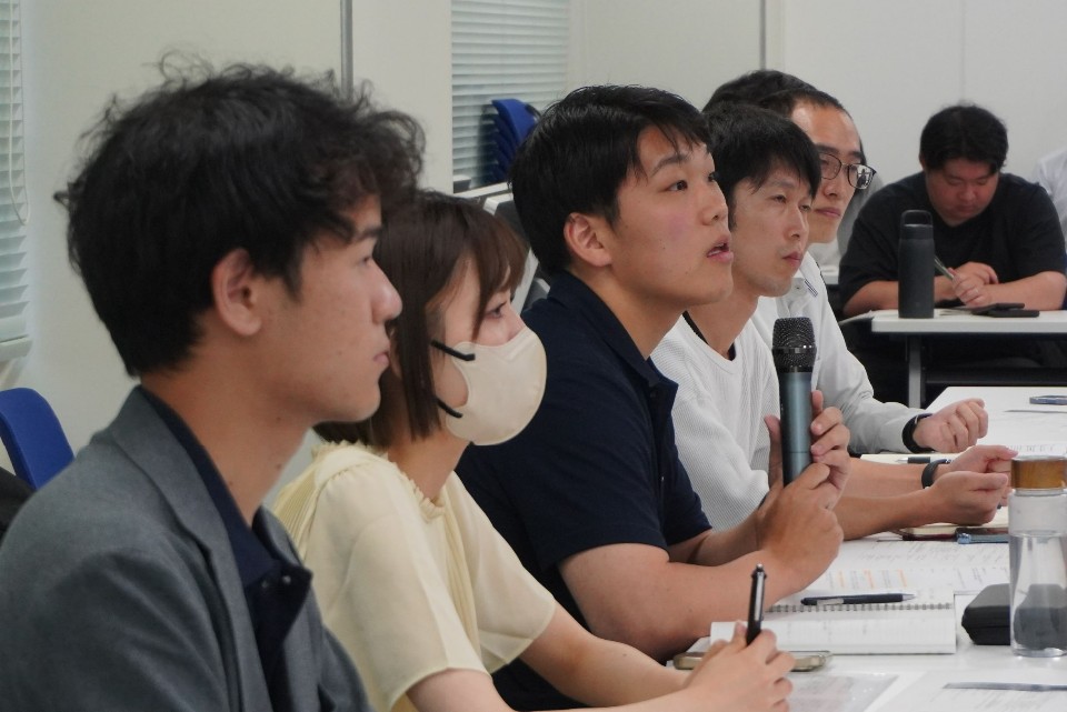 研修会場で横一列に座る男女が真剣な表情で前方を注視しており、中央の紺色のポロシャツを着た男性がマイクを手に持ちながら発言したり質問したりしている、セミナーの風景を捉えた写真